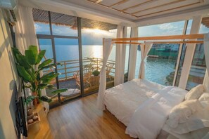 Une chambre avec une grande fenêtre donnant sur un plan d’eau, un lit, une plante et un petit coin salon avec une chaise et une table.