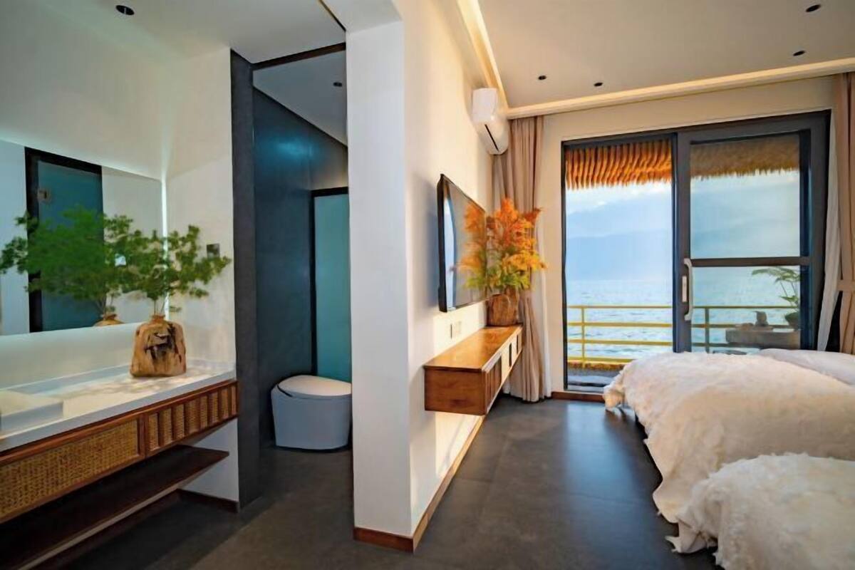 Habitación de hotel moderna con una cama grande, un escritorio con una planta, un espejo y vista al mar a través de un ventanal amplio.