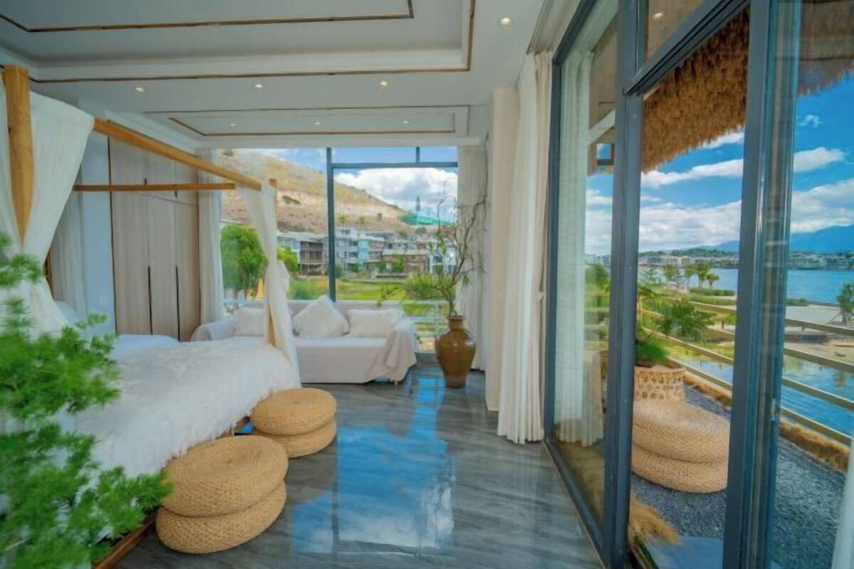 Un hotel moderno con un amplio ventanal, un sofá blanco, una cama con ropa de cama blanca y vistas a una masa de agua y colinas.