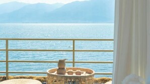 Ein Zimmer mit Blick auf ein ruhiges Meer und ferne Berge, ein Fenster mit Vorhang und ein kleiner Tisch mit einem dekorativen Gegenstand.