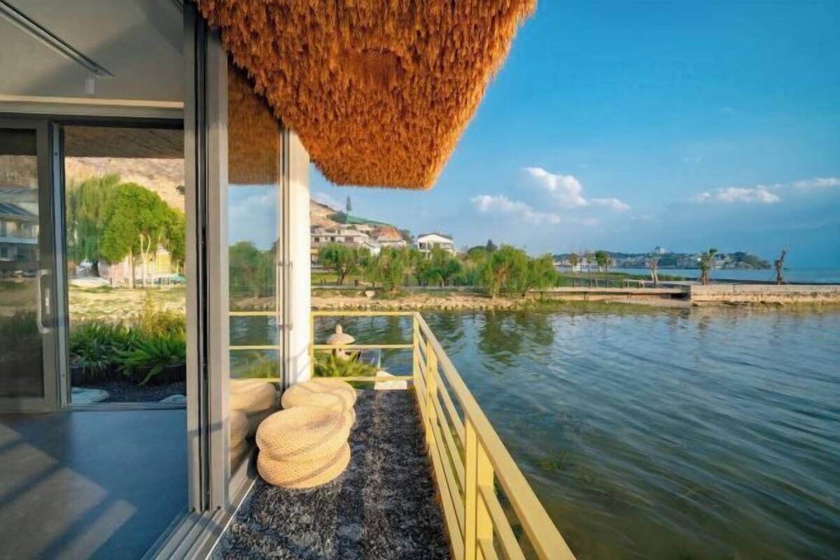 Un edificio moderno con fachada de vidrio, techo de paja y vistas a un lago tranquilo con un muelle.