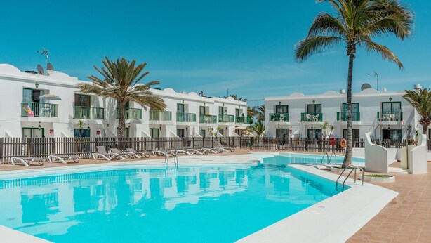 Une piscine entourée de bâtiments blancs dotés de balcons, de palmiers et de chaises longues.