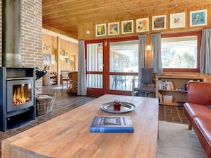 A cozy living room with a fireplace, a wooden table with a book, a leather sofa, and a large window with curtains. - 6 Person Holiday Home in Blokhus (Blokhus)