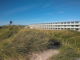 A modern building with a flat roof and multiple windows, surrounded by grassy dunes.