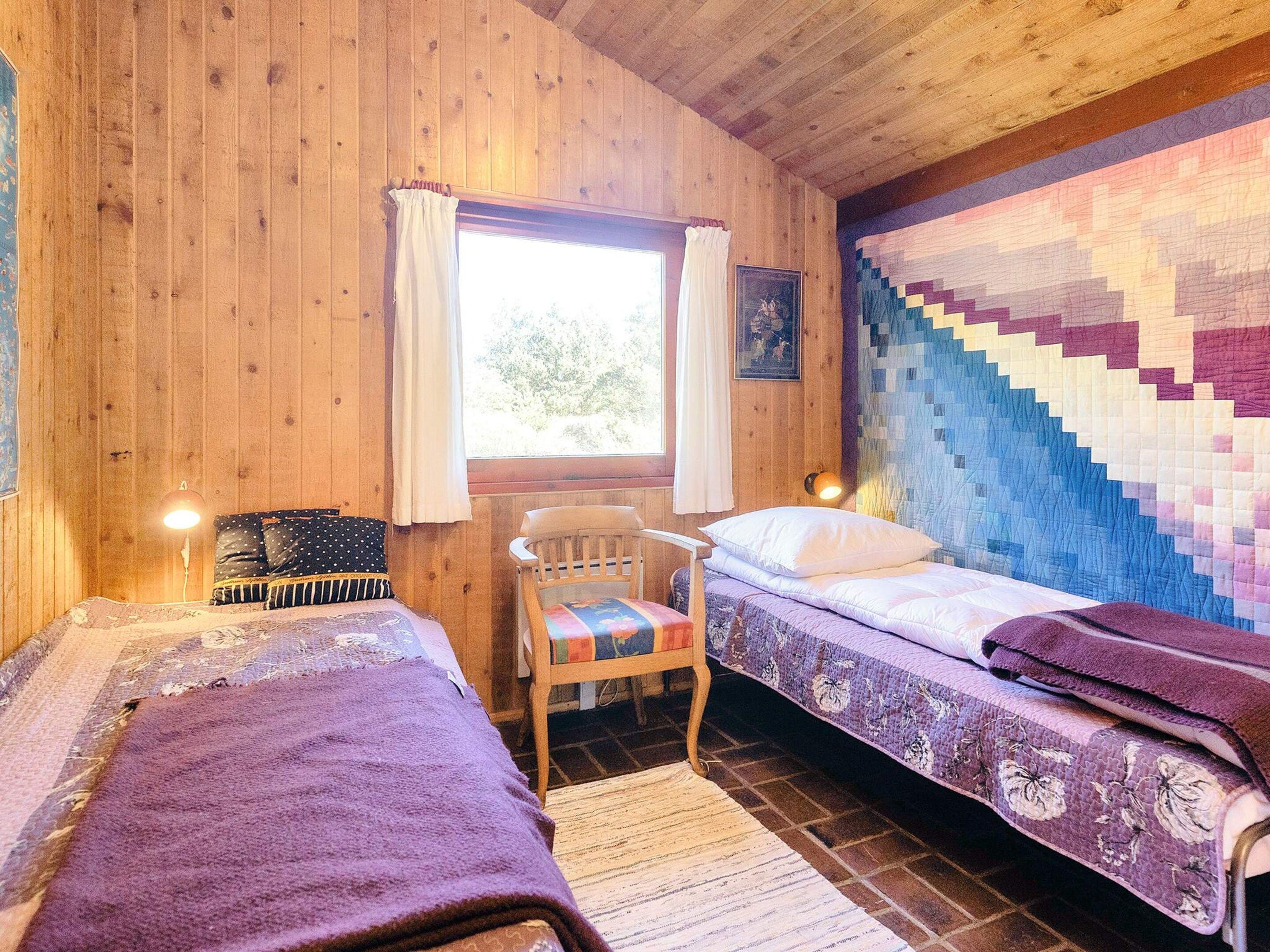 Habitació de cabana amb dos llits, una cadira de fusta, una finestra amb cortines i un tapís de paret acolxat.