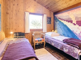 A cozy cabin room with two beds, a wooden chair, a window with curtains, and a quilted wall hanging.