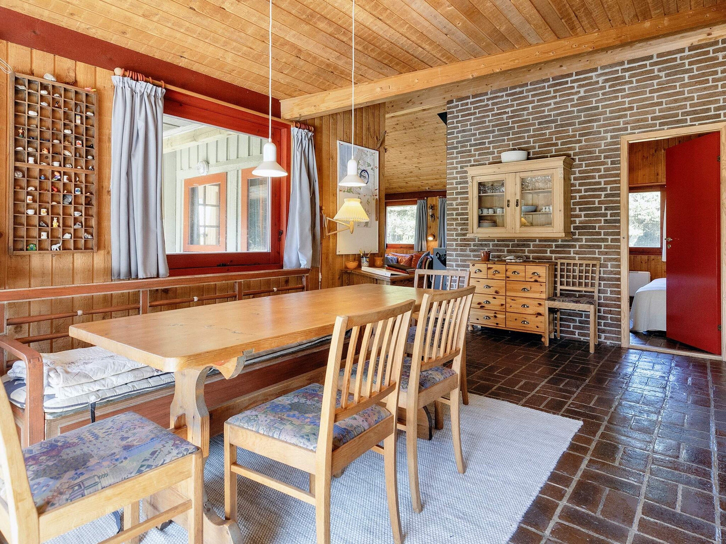 Een houten interieur met een lange eettafel, stoelen, een bakstenen muur en een rode deur.