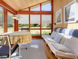 A room with a wooden ceiling, a round table with a patterned tablecloth, a sofa with patterned cushions, and large windows with white curtains.