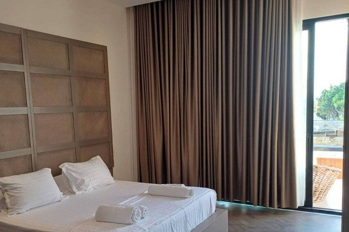 A modern hotel room with a large bed, a headboard with a geometric pattern, and a view of the outdoors through a large window with curtains.