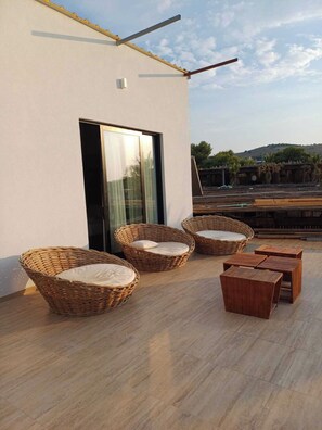 Una terraza con sillas de mimbre y mesas de madera, un ventanal y vistas del entorno.