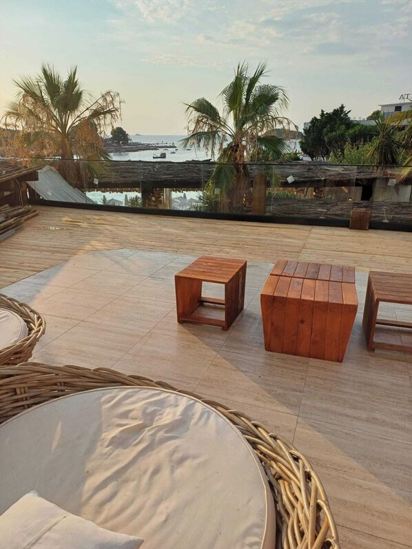 Terraza de madera con dos mesas de madera y una silla de mimbre, con vistas a una playa con palmeras y una estructura de techo de paja.