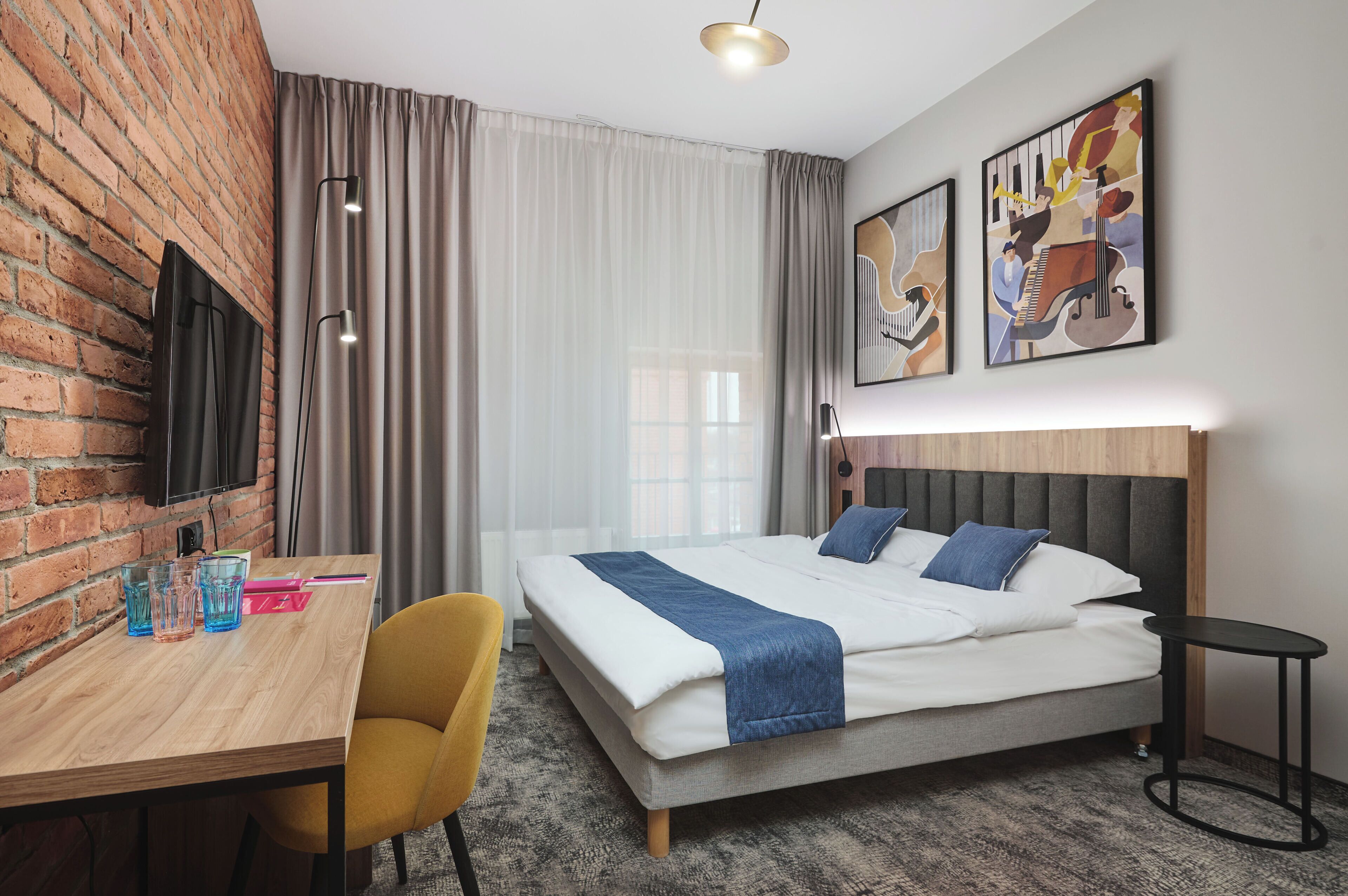 A hotel room with a brick wall, a bed with a headboard, a desk with a chair, and artwork on the wall.