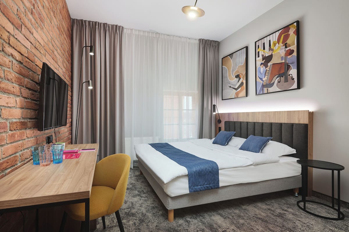 Une chambre d’hôtel comprenant un lit, un bureau, une chaise, un téléviseur fixé au mur et des œuvres d’art accrochées au mur.