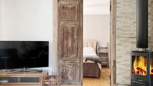 Uma sala de estar acolhedora, com uma porta de madeira que dá para um quarto, uma televisão, uma lareira e uma pequena mesa de madeira.