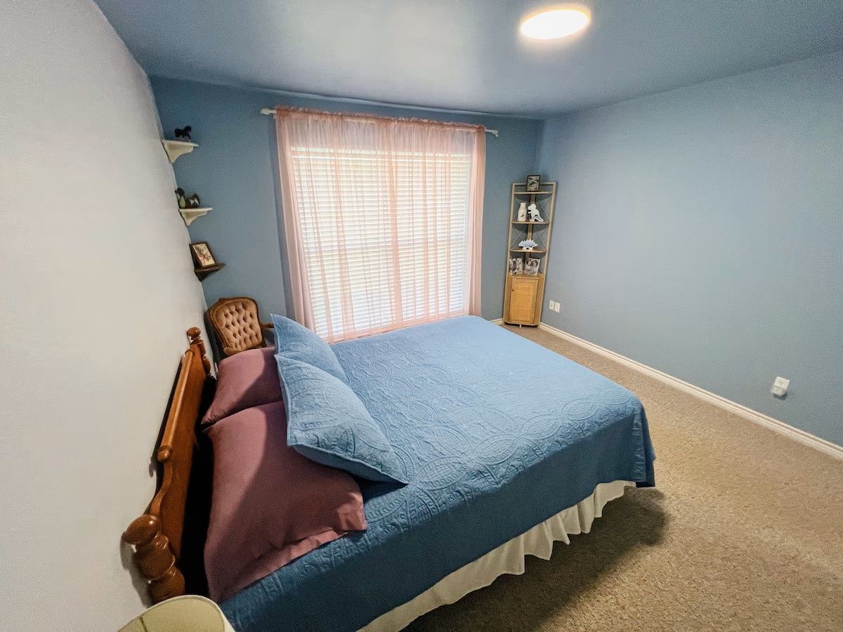 Un dormitorio con cama, ventana con persianas, mesita de noche y estante con objetos decorativos.