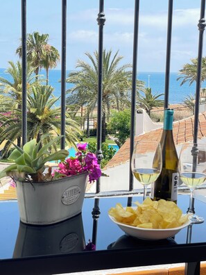 Un balcón con una mesa que incluye una botella de vino, un cuenco de papas fritas y una maceta con flores rosas.