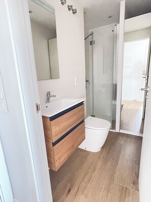Un baño moderno con tocador de madera, un lavamanos blanco, un inodoro y una cabina de ducha de vidrio.