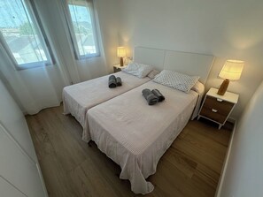 Habitación doble con dos camas, cada una con una almohada y una toalla enrollada. Hay mesitas de noche con lámparas y una ventana con cortinas.