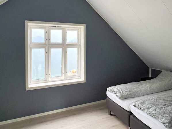 Un dormitorio con techo inclinado, una ventana con marcos blancos y una cama con manta gris.