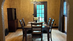 A dining room with a wooden table and chairs, a chandelier, and a window with curtains. - Historic Twin Tudors Inn Southeast Room (KP1400SE) (Sioux Falls)