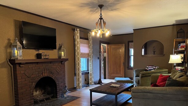A living room with a fireplace, a flat-screen TV mounted above it, a sofa, a coffee table, and a chandelier. - Historic Twin Tudors Inn Southeast Room (KP1400SE) (Sioux Falls)