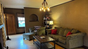 A cozy living room with a wooden coffee table, a green sofa with red cushions, a striped throw blanket, and a bookshelf. - Historic Twin Tudors Inn Southeast Room (KP1400SE) (Sioux Falls)