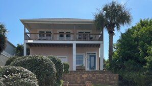 Une maison à deux étages avec un porche à l’avant, un escalier en pierre menant à l’entrée et un palmier en arrière-plan.