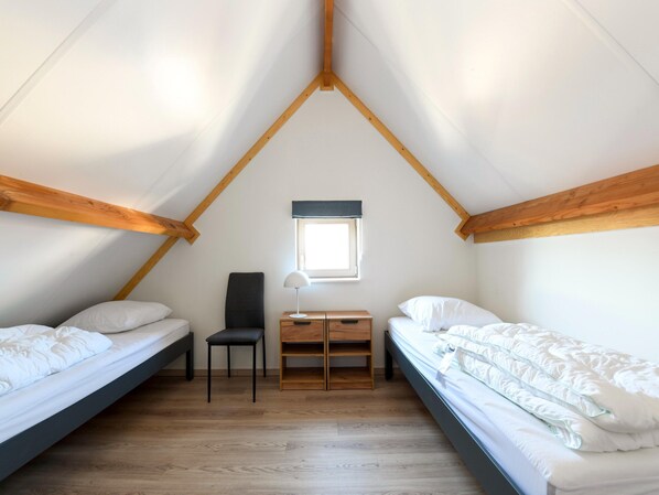Une petite chambre mansardée de forme triangulaire, avec deux lits, une chaise, un bureau et une fenêtre.