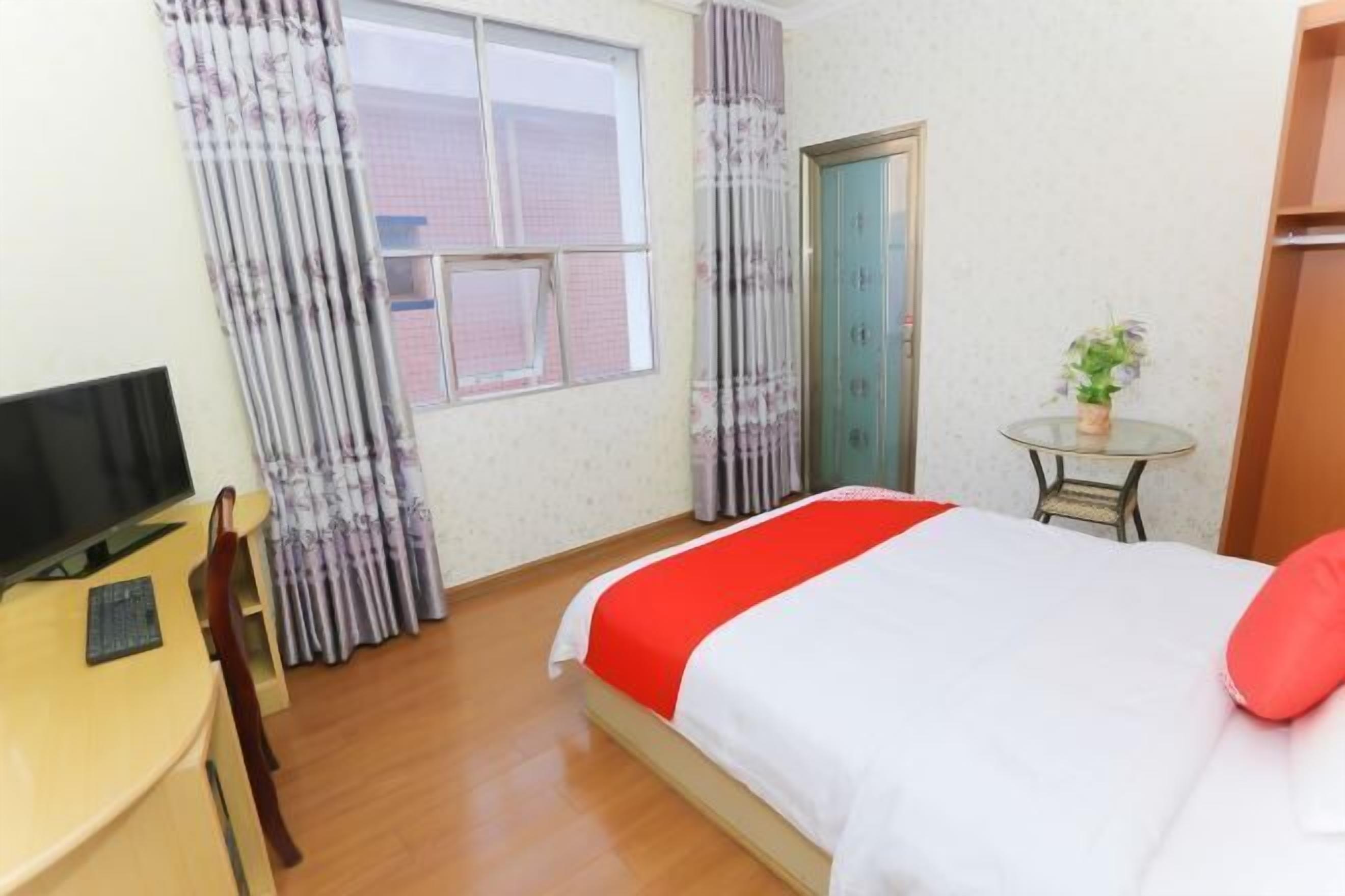 A hotel room with a bed, a TV, a desk, a chair, a small table with a plant, and a window with curtains.