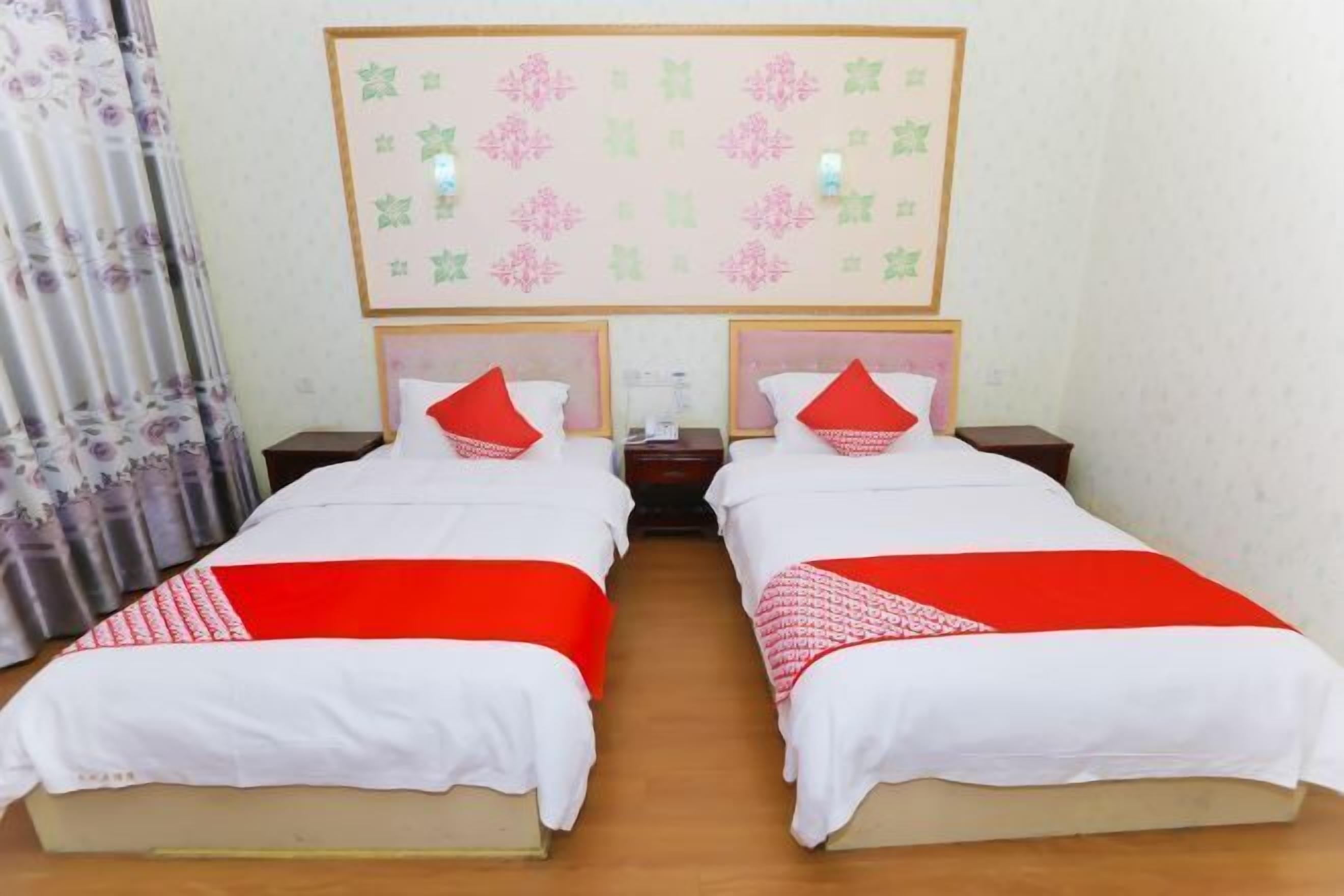 A hotel room with two beds, each with a red and white bedspread, a headboard with a floral pattern, and a wall-mounted light fixture.