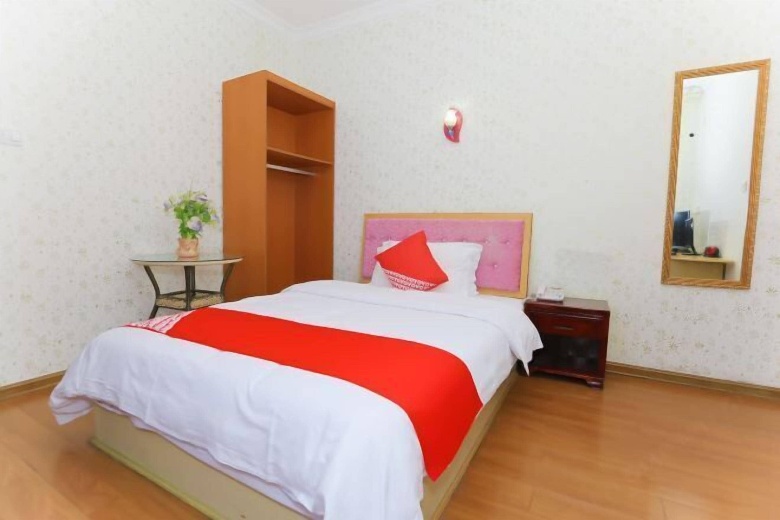 A hotel room with a bed, a bedside table, a small round table with a plant, a mirror, and a wardrobe.