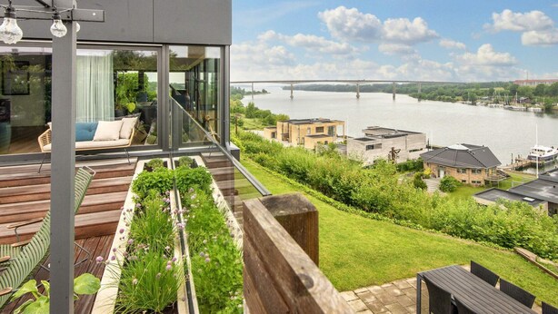 Une maison moderne dotée d’un balcon donnant sur une rivière, un pont et un port de plaisance.