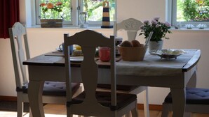Een eettafel met stoelen, een vaas met bloemen, een schaal brood en een raam met uitzicht op groen.