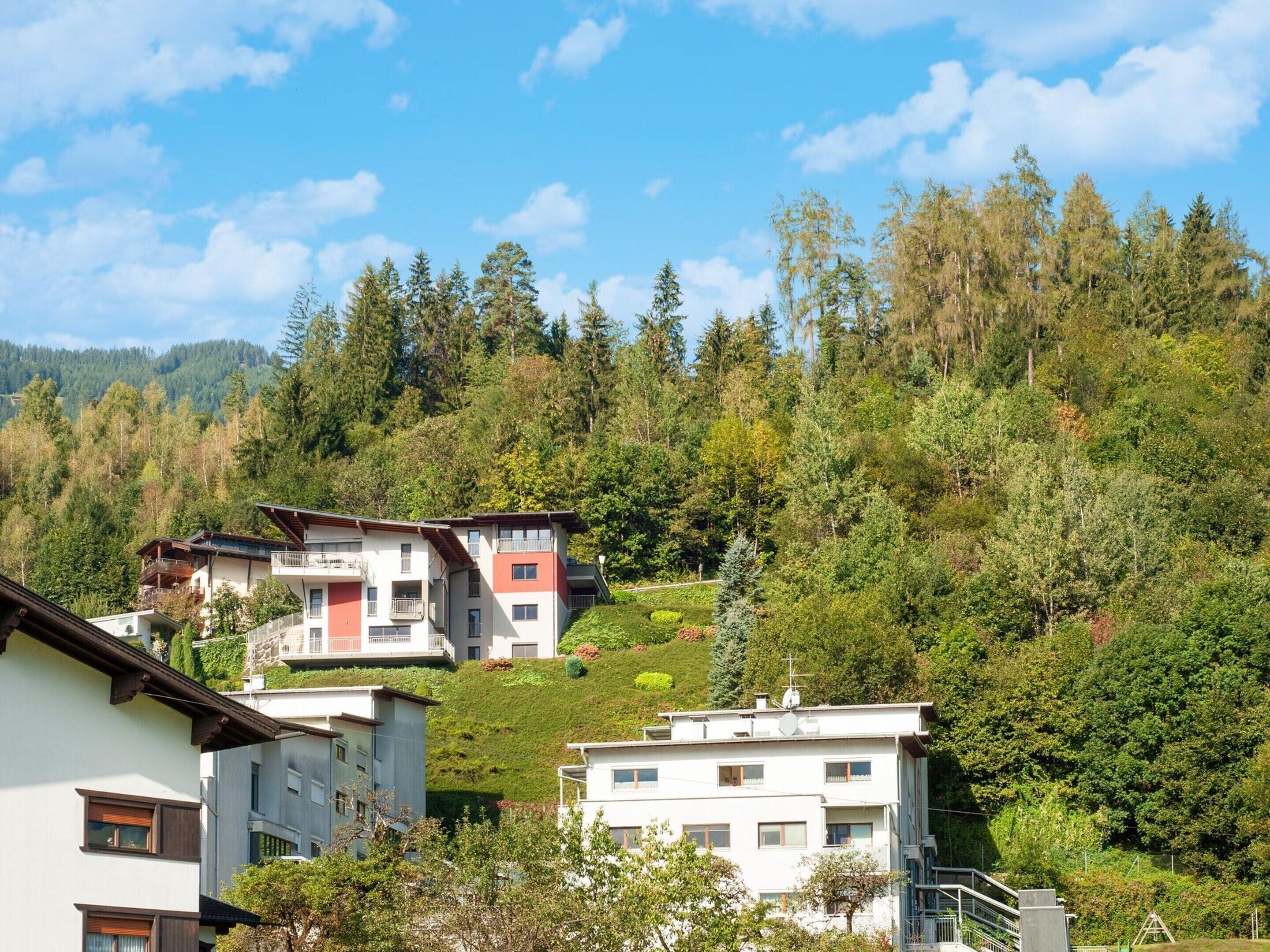 Una zona residencial con edificios de varios pisos, algunos con balcones, con un fondo de bosque denso y colinas.