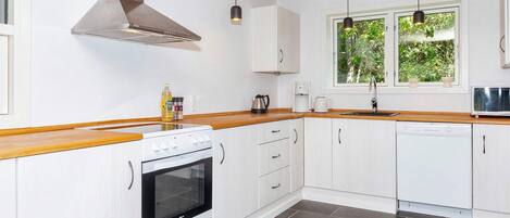 A modern kitchen with white cabinetry, a wooden countertop, a stainless steel range hood, a built-in oven, a dishwasher, and a window with a view of greenery.