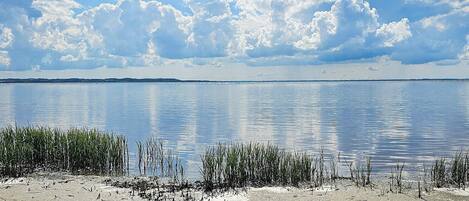 A calm lake with a sandy shore and tall grasses in the foreground, under a partly cloudy sky.