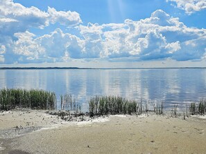 Un lac paisible, avec une rive sablonneuse et de hautes herbes au premier plan, sous un ciel partiellement nuageux.