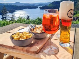 Una mesa con un tazón de papas fritas, un vaso de bebida roja y un vaso alto de cerveza con un logotipo, frente a una vista panorámica de un lago.
