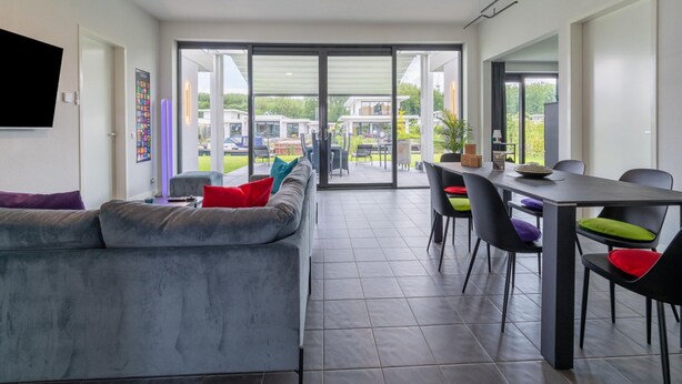 A modern living room with a grey sofa, a dining table with chairs, and a large sliding glass door leading to an outdoor patio. - Villa With Covered Terrace and Jetty on the Water (Zeewolde)