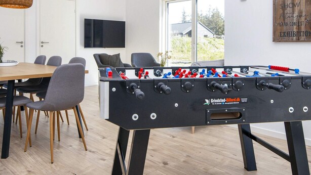 A modern room with a foosball table, a dining area with chairs and a table, a TV mounted on the wall, and a window with a view of the outdoors.