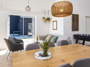 A modern living room with a wooden dining table, grey sofas, a foosball table, and a large window overlooking a dark exterior wall.