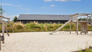 A sandy beach area with a playground featuring a slide and swings, and a building with a tiled roof in the background.
