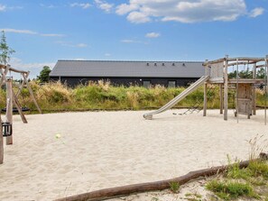A sandy beach area with a playground featuring a slide and swings, and a building with a tiled roof in the background. - 8 Person Holiday Home in Romo (Rømø)