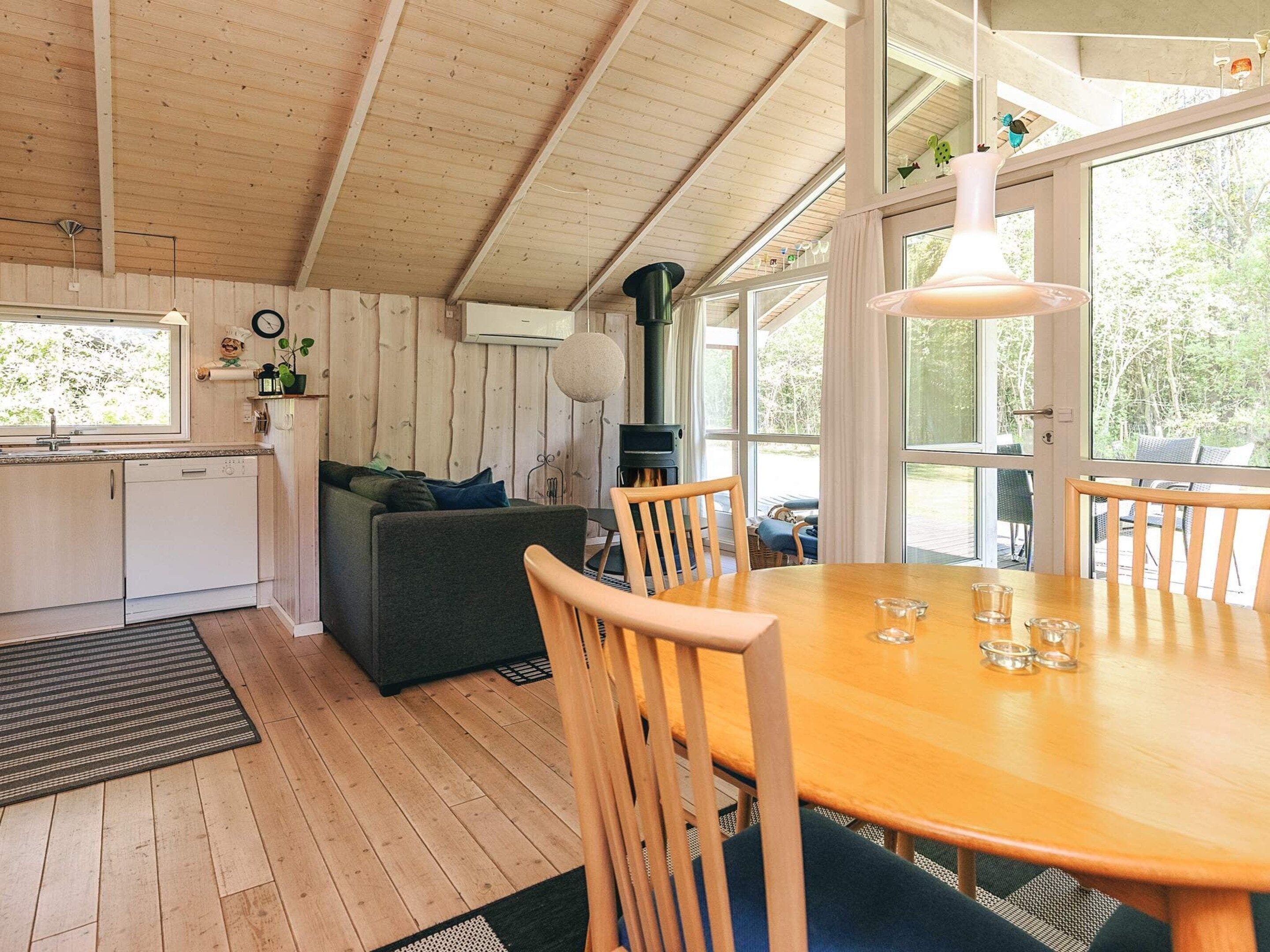 A cozy cabin interior with a wooden ceiling, a dining table with chairs, a sofa, and a kitchen area with a dishwasher.