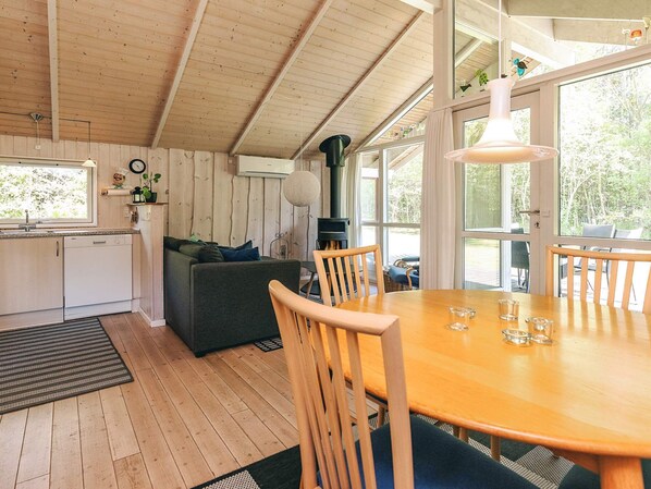 A cozy cabin interior with a wooden ceiling, a dining table with chairs, a sofa, and a kitchen area with a dishwasher.