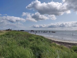 A coastal landscape with grassy dunes, a pier extending into the sea, and a small building on the left.