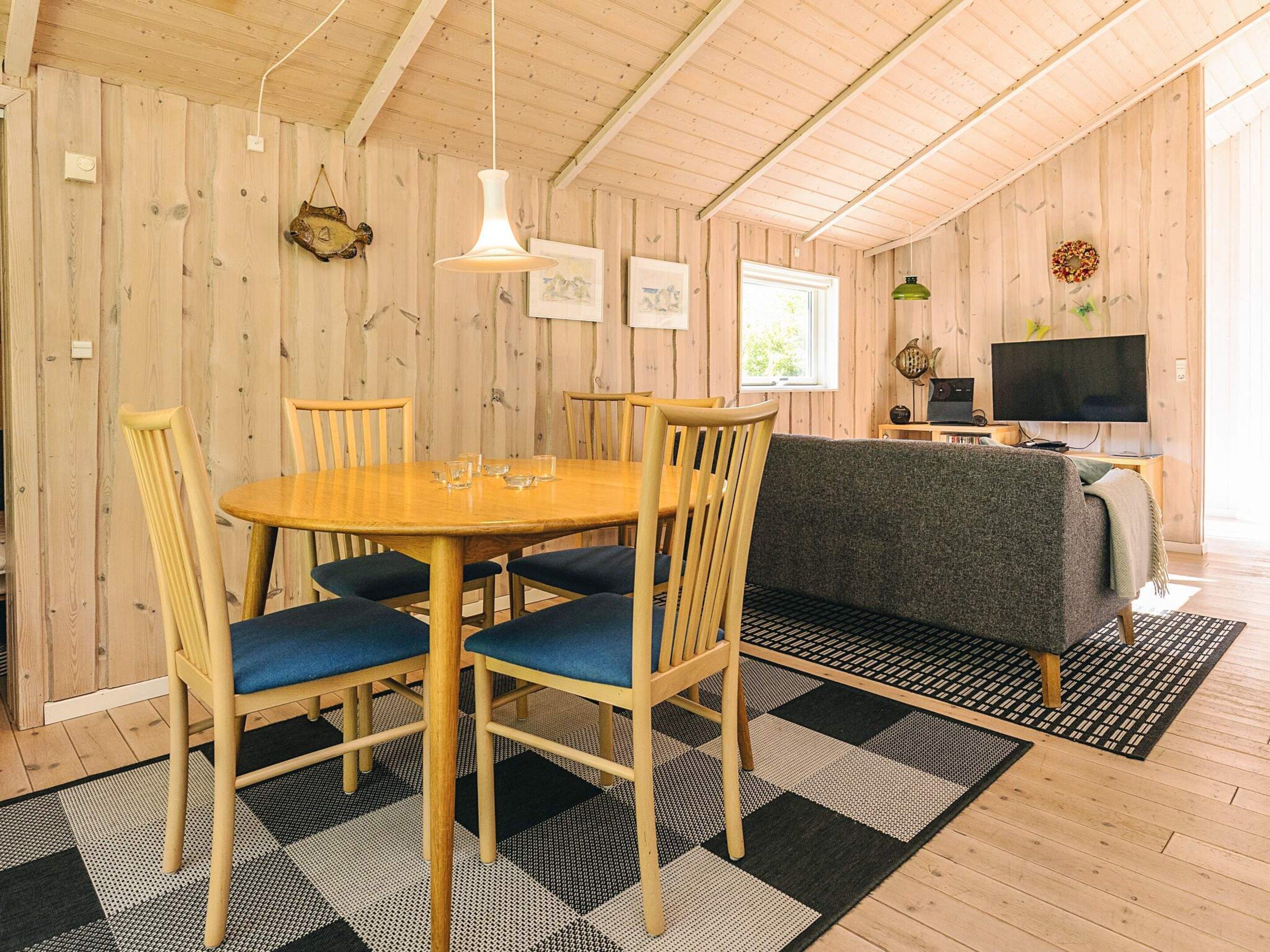Un interior de madera con una mesa redonda para comer, sillas con cojines azules, un sofá, un televisor y una alfombra estampada.