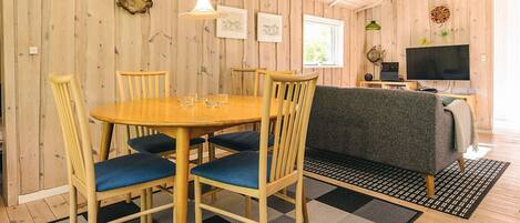 Un interior de madera con una mesa redonda para comer, sillas con cojines azules, un sofá, un televisor y una alfombra estampada.