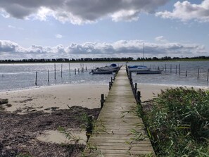 Eine hölzerne Pier führt zu Booten auf ruhigem Wasser, umgeben von Vegetation und einem bewölkten Himmel.
