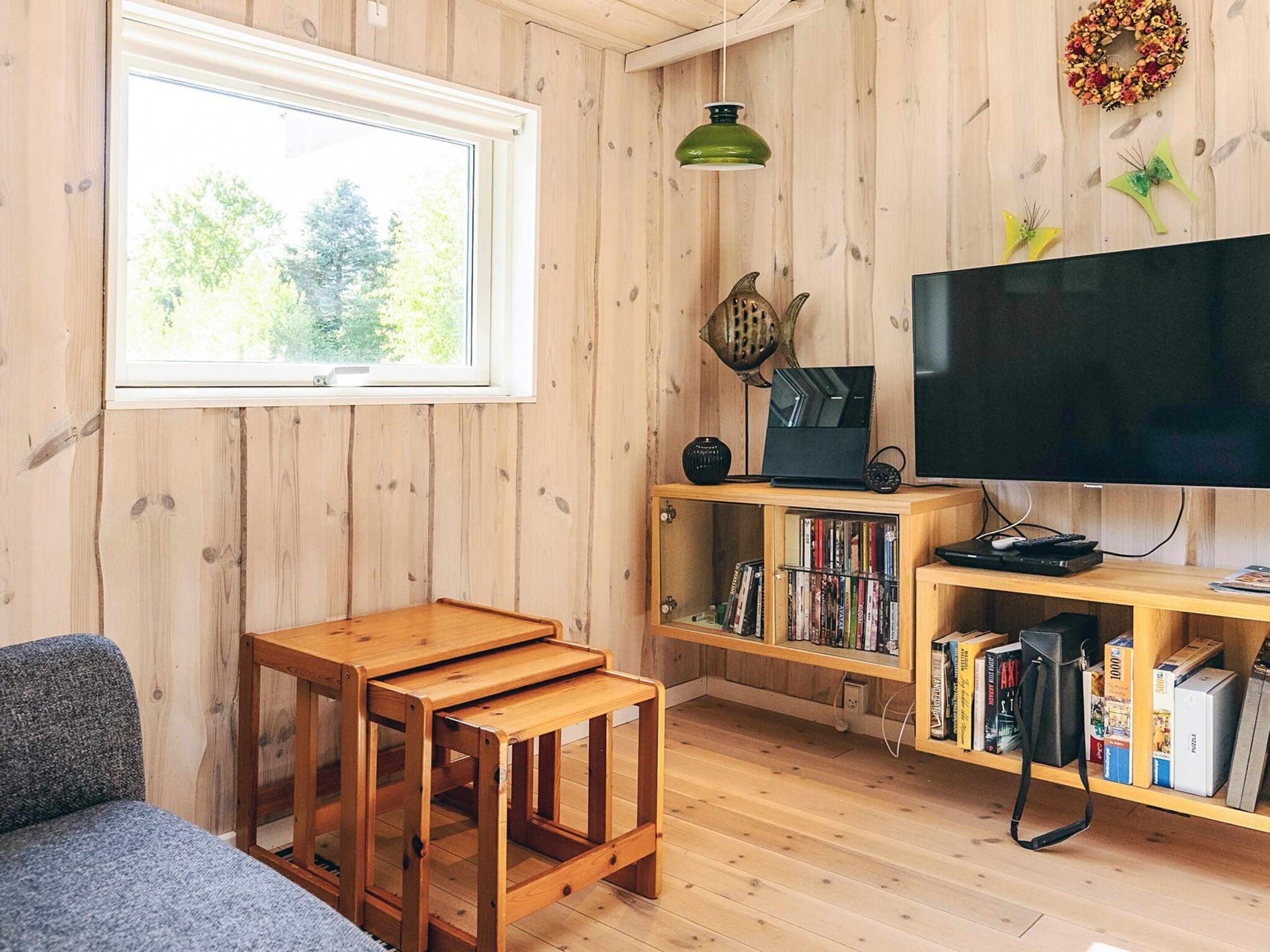 A cozy living room with a wooden floor, a large window, a flat-screen TV, a bookshelf, and a small wooden table.
